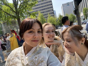Three USAC students wearing traditional Korean hanbok take a selfie outdoors on a sunny street in Seoul, South Korea, with trees, buildings, and other people in the background.