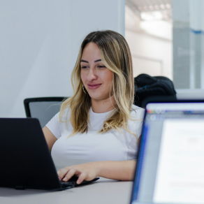 Eine blonde, l&auml;chelnde Frau sitz in einem Computerraum und arbeitet auf einem Laptop