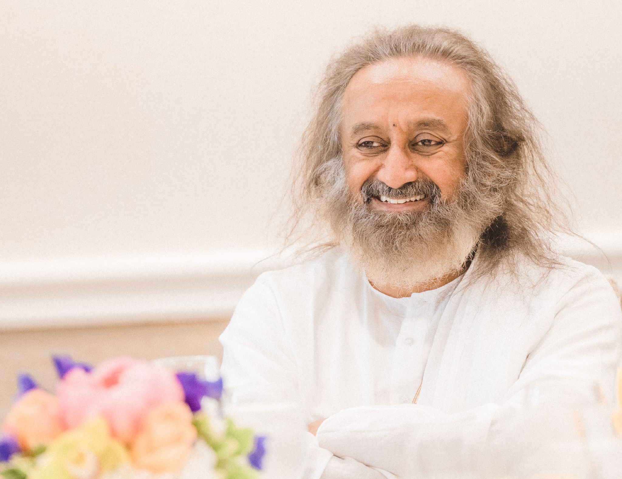 Retrato de Sri Sri Ravi Shankar sonriente vestido de blanco con flores de colores y luz cálida